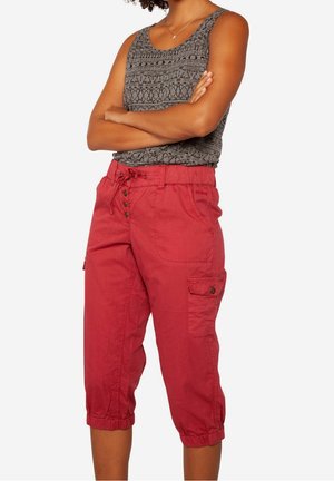 Frau trägt ärmelloses graues Oberteil mit Muster und rote Cargohose in Mid-Calf-Länge mit Knopf- und Kordelzugbund, steht mit verschränkten Armen.