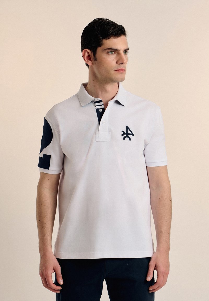 Camisa polo blanca con detalles en azul marino, con un cuello, tres botones y un logotipo en el pecho. Las mangas tienen un diseño en contraste.