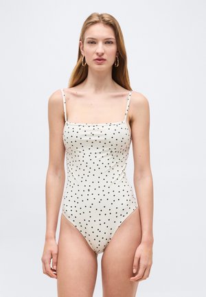 Młoda kobieta w białym jednoczęściowym kostiumie kąpielowym z cienkimi ramiączkami i czarnymi kropkami, stojąca na jednolitym jasnym tle.