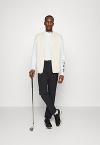 Quilted cream vest over a light blue long-sleeve shirt, paired with black pants and black shoes, holding a golf club.