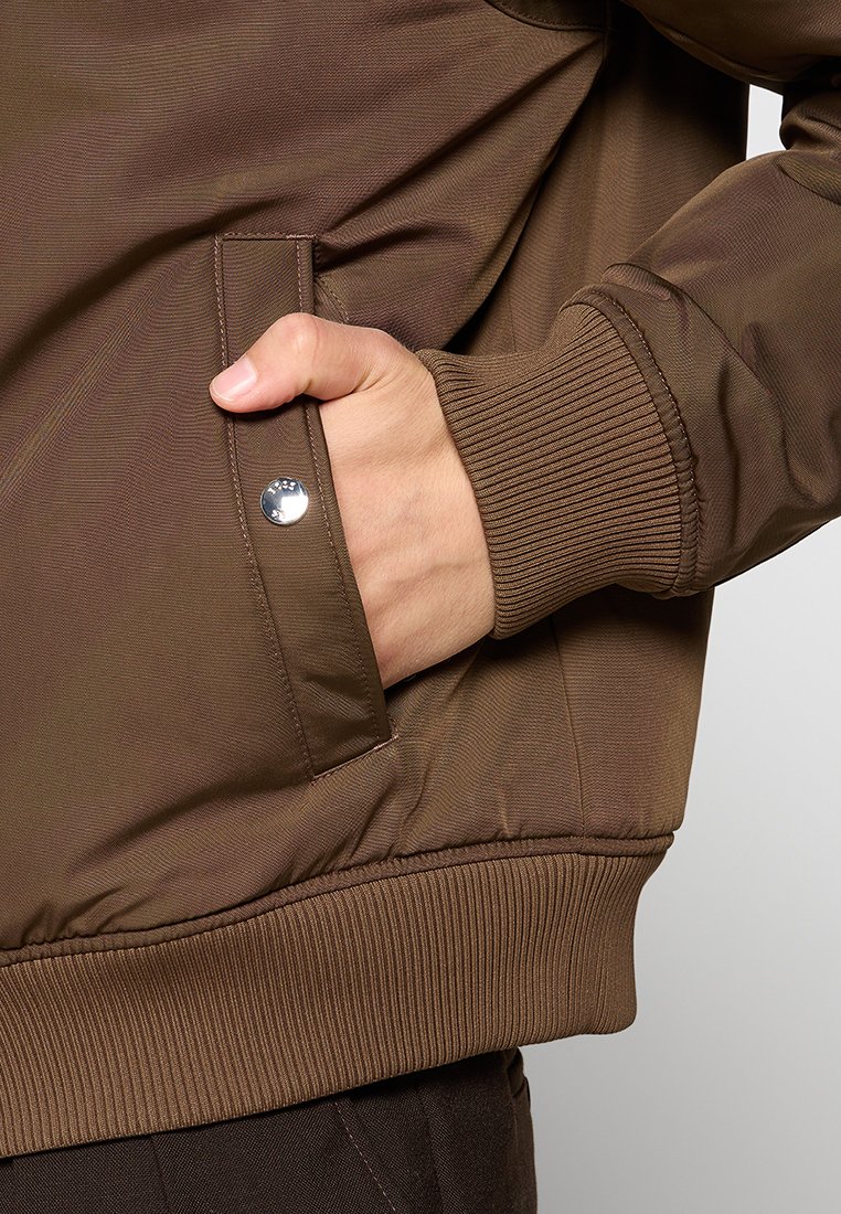 Veste bombardier marron avec des poignets côtelés, des poches latérales et une texture lisse. Présente un détail à bouton pression argenté près de la poche.