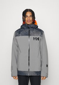 Veste imperméable grise avec capuche, présentant des accents noirs, une doublure orange et un logo sur la poitrine. Manches longues et coupe décontractée.