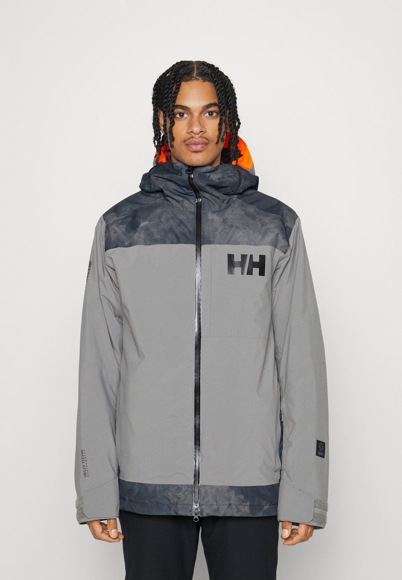 Veste imperméable grise avec capuche, présentant des accents noirs, une doublure orange et un logo sur la poitrine. Manches longues et coupe décontractée.