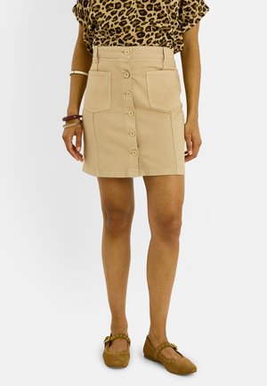 Mujer con falda beige con botones delanteros y bolsillos frontales, top con estampado de leopardo y bailarinas Mary Jane marrones con tachuelas sobre fondo blanco.