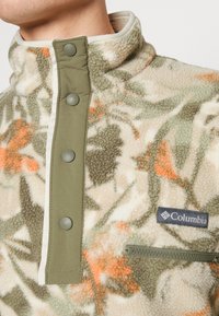 Pullover in felpa beige con motivi floreali verdi, dotato di colletto alto, bottoni a scatto e tasca sul petto con zip. Logo Columbia in evidenza.