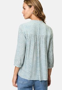 Helle blaue Bluse mit floralem Muster, 3/4-Ärmeln, rundem Halsausschnitt und gerafftem Rückendetail. Kombiniert mit hellblauen Jeans.
