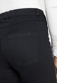 Pantaloni in denim neri con una tasca posteriore con dettagli cuciti e un piccolo logo del brand, accompagnati da un top bianco testurizzato.