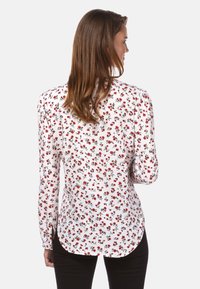 Weiße Bluse mit kleinem roten Blumenmuster, langen Ärmeln und taillierter Form. Rundsaum und leicht geraffte Schulterdetails.