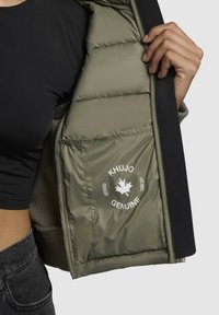 Jachetă căptușită cu un interior verde măsliniu, având un patch cu logo și un strat exterior negru. Interiorul prezintă un model texturat, matlasat.
