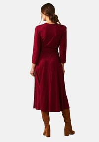 En kvinna sedd bakifrån bär en lång bordeauxfärgad klänning med trekvartsärmar, kombinerad med höga bruna stövlar, stylad i en minimalistisk miljö.