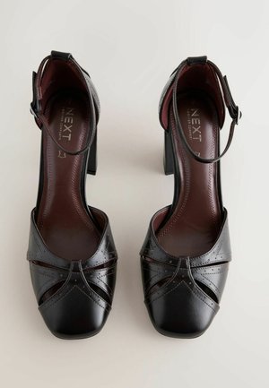 Mustat nahkakengät korkeilla koroilla, joissa on ristikkäinen punottu kuvio, neliönmuotoiset kärjet ja nilkkaremmit. Sisäpuolella on viininpunainen vuori ja pehmusteet.