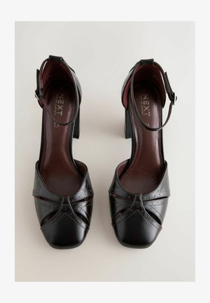 Mustat nahkakengät korkeilla koroilla, joissa on ristikkäinen punottu kuvio, neliönmuotoiset kärjet ja nilkkaremmit. Sisäpuolella on viininpunainen vuori ja pehmusteet.