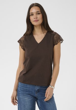 Žena s dlhými hnedými vlasmi, oblečená v tmavohnedom tričku s výstrihom do V a čipkovanými krátkymi rukávmi, a modrých džínsoch, na jednoduchom pozadí.