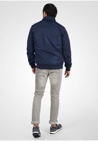 Navy blue bomber jacket with a stand collar, elastic cuffs, and hem; paired with light grey pants. Casual footwear completes the look.