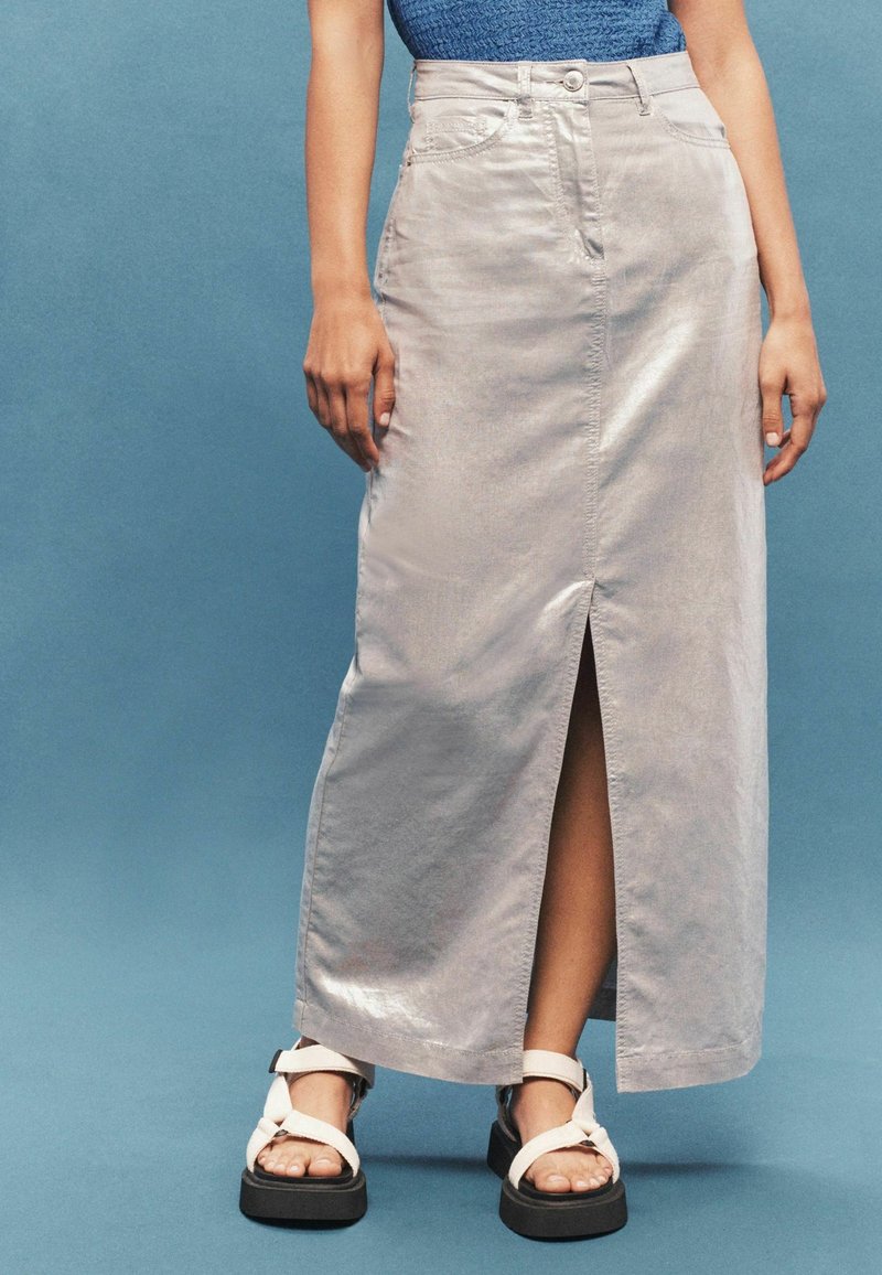 Femme portant une longue jupe gris clair avec une fente à l'avant, des sandales compensées blanches et un top bleu sans manches sur fond bleu.
