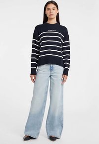 Maglione a righe navy con linee orizzontali bianche, orlo a coste e scollo rotondo, abbinato a jeans a gamba larga di colore azzurro chiaro. Scarpe nere.