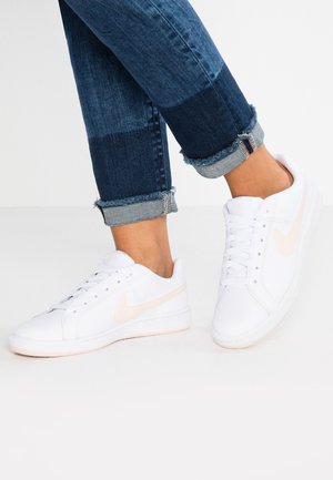 Hvide atletiksneakers med en lys ferskenfarvet Nike-swoosh, tekstureret læderoverdel og en gummisål. Båret med rullede denimjeans.