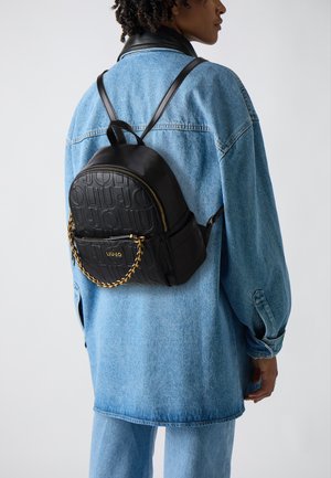 Zaino nero con motivo a logo in rilievo, dettaglio della catena dorata e chiusura con zip, indossato sopra una giacca di denim blu chiaro.