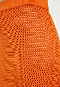 Oranje gaasstof met een gestructureerd patroon en kleine gaatjes, gecombineerd met een solide oranje band aan de bovenkant voor een veilige pasvorm.