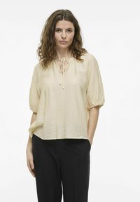 Top corto de manga corta en color beige claro con una tela texturizada, escote redondo y detalle de lazo en el frente. Combinado con pantalones negros.