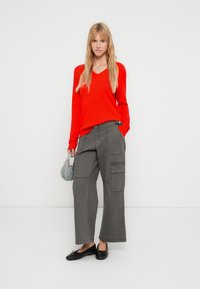 Κόκκινο πουλόβερ με V λαιμόκοψη, γκρι φαρδιά παντελόνια cargo με πλαϊνές τσέπες και μαύρα μπαλαρίνα παπούτσια. Το μοντέλο κρατά μια μικρή γκρι τσάντα. Το φόντο είναι απλό λευκό.