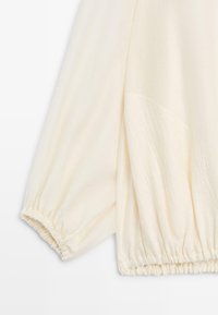 Mouw van crèmekleurige blouse met geplooide elastische manchetten en subtiele, gestructureerde stofdetail.