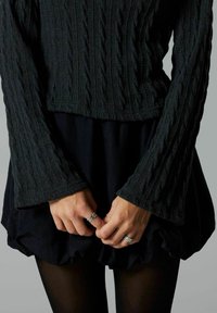 Maglione nero a maglia con maniche svasate abbinato a una gonna scura a balze. Le mani indossano anelli in argento e collant scuri. Design semplice e strutturato.