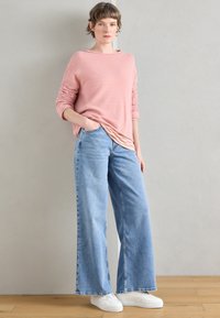 Camisola de malha rosa claro com mangas compridas por cima de uma t-shirt às riscas, combinada com jeans azul claro de pernas largas e ténis brancos.