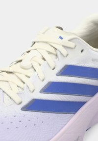 Λευκό αθλητικό παπούτσι με ανοιχτό μπλε λεπτομέρειες, ύφασμα με υφή, επίπεδα κορδόνια και τρεις μπλε ρίγες στο πλάι. Λεπτομέρεια στη σόλα σε ανοιχτό μωβ χρώμα.