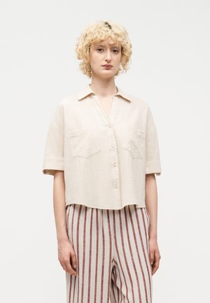 Νεαρό άτομο με σγουρά ξανθά μαλλιά φορά χαλαρό, εκρού πουκάμισο κοντού μανικιού με κουμπιά και μπεζ παντελόνι με ψηλή μέση και κάθετες καφέ ρίγες.