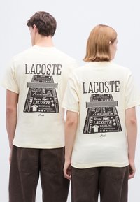 Βαμβακερό μπλουζάκι σε κρεμ απόχρωση με μαύρο σχέδιο στο πίσω μέρος, που απεικονίζει κείμενο και εικόνες σχετικές με το τένις. Οι άκρες είναι ραμμένες προσεκτικά.