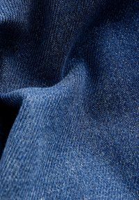 Gros plan d’un tissu en denim bleu foncé texturé avec un tissage visible et des plis naturels créant des variations de lumière et d’ombre.