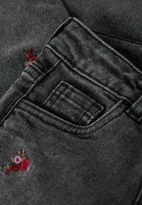 Donkergrijze denim stof met een klein geborduurd bloemenmotief op de voorzak, met een rechte snit en zichtbare stiksels.