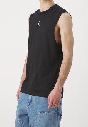 Modèle masculin portant un débardeur noir Jordan et un short cargo en jean bleu clair, debout devant un fond blanc uni.