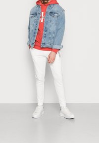 Personne portant un sweat à capuche rouge, une veste en denim bleu clair, un pantalon de jogging blanc avec logo et des baskets blanches, se tenant devant un fond blanc uni.