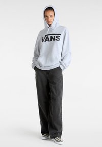 Felpa grigia con logo "VANS" nero, cappuccio con cordini e tasca a marsupio. Indossata con pantaloni larghi scuri e scarpe slip-on a scacchi.
