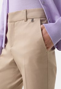 Beige tailored pants with a smooth texture, featuring a front pocket and a subtle metal accent near the waistband. Paired with a lavender knit sweater.