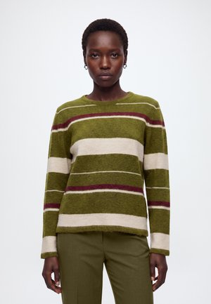 Femme aux cheveux courts porte un pull vert rayé avec des bandes blanches et bordeaux ainsi qu'un pantalon vert olive, debout devant un fond blanc uni.