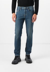 Jeans de un azul oscuro con un corte slim, lavado medio y un ligero desgaste. Presentan cinco bolsillos y un cierre de cremallera, combinados con zapatillas deportivas negras.