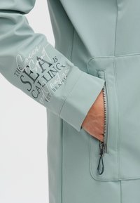Light blue jacket featuring a zippered pocket, a smooth texture, and printed text on the sleeve. Zipper pull has a rubber accent.