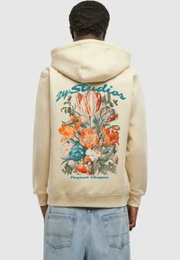 Sudadera crema con capucha que presenta un gran estampado floral en colores vibrantes y el texto "24/7 Studios" con un acento azul.