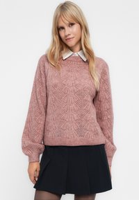 Giovane donna con lunghi capelli biondi che indossa un maglione lavorato a maglia rosa con motivo, una camicia bianca con colletto, una gonna plissettata nera e collant trasparenti.