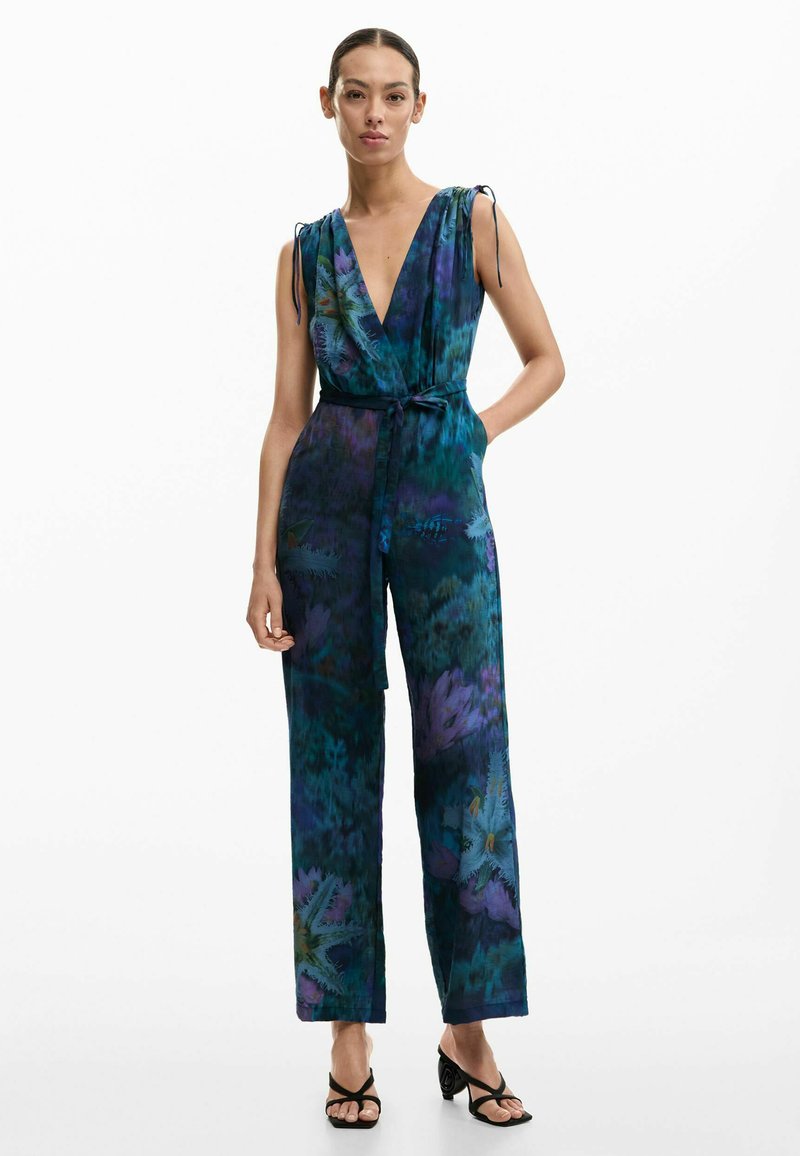 Kvinde iført ærmeløs jumpsuit med dyb V-hals og bælte, med blå og lilla blomsterprint, kombineret med sorte sandaler med hæl og stropper.