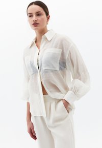 Biała, przezroczysta koszula zapinana na guziki z dwoma przednimi kieszeniami, długimi rękawami i luźnym krojem. Materiał wydaje się lekki i przezroczysty.
