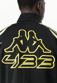 Black jacket featuring yellow embroidered graphic of two seated figures and the number "423." Includes white accent details along the seams.