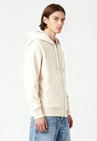 Felpa con zip beige realizzata in tessuto morbido, con tasca frontale, cappuccio con cordino e polsini a costine. Indossata con jeans azzurri chiari.