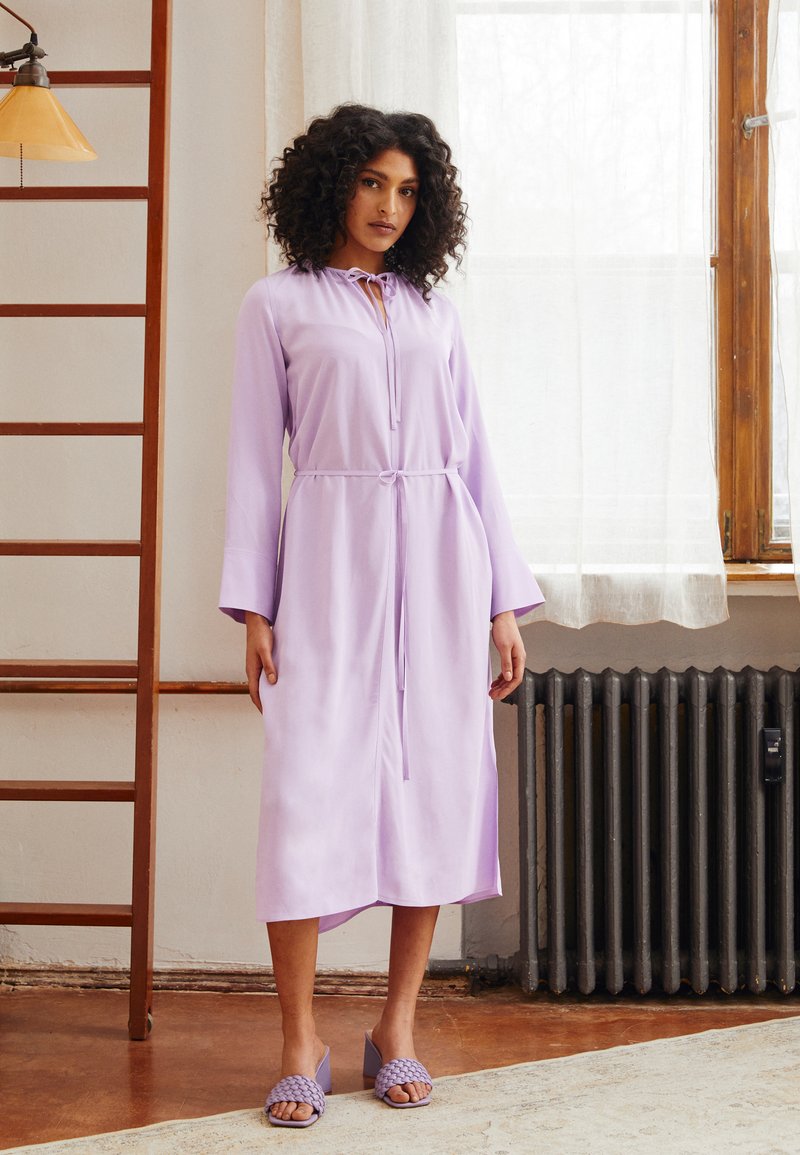 Vestido longo lilás com fecho de laço no pescoço, cintura ajustada e mangas com efeito ''bell''. Combinado com sandálias roxas de slide trançadas.