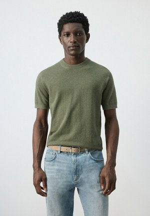 Ung mand står mod en ensartet baggrund iført en olivengrøn kortærmet sweater, lyseblå jeans og beige bælte.