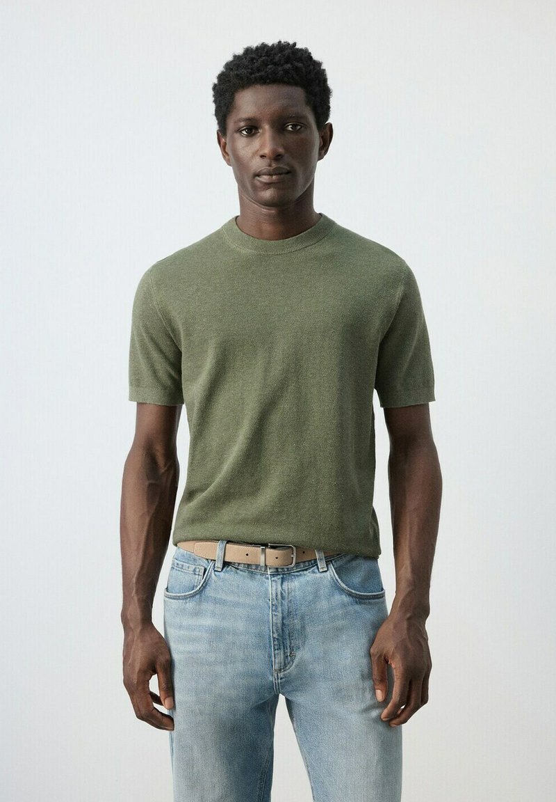 Mladý muž stojaci pred jednoduchým pozadím, má na sebe olivovo zelený krátky sveter, svetlomodré rifle a béžový opasok.