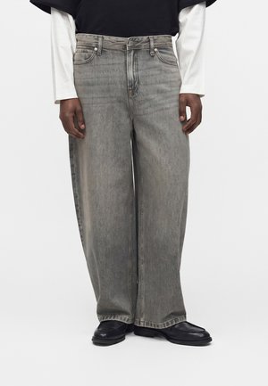 Osoba nosiaca voľné sivé vyblednuté rifle s čiernymi topánkami a čiernym topom cez bielu košeľu s dlhým rukávom stojaca pred bielym pozadím.